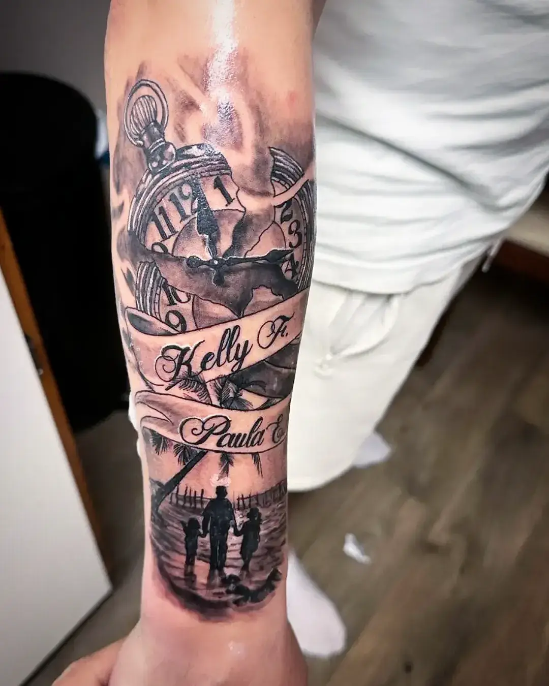 Black and grey memorial forearm sleeve with pocket watch, family beach silhouette, and "Kelly F" / "Paula" banners by Fresiato at Shadow Studio Charlotte