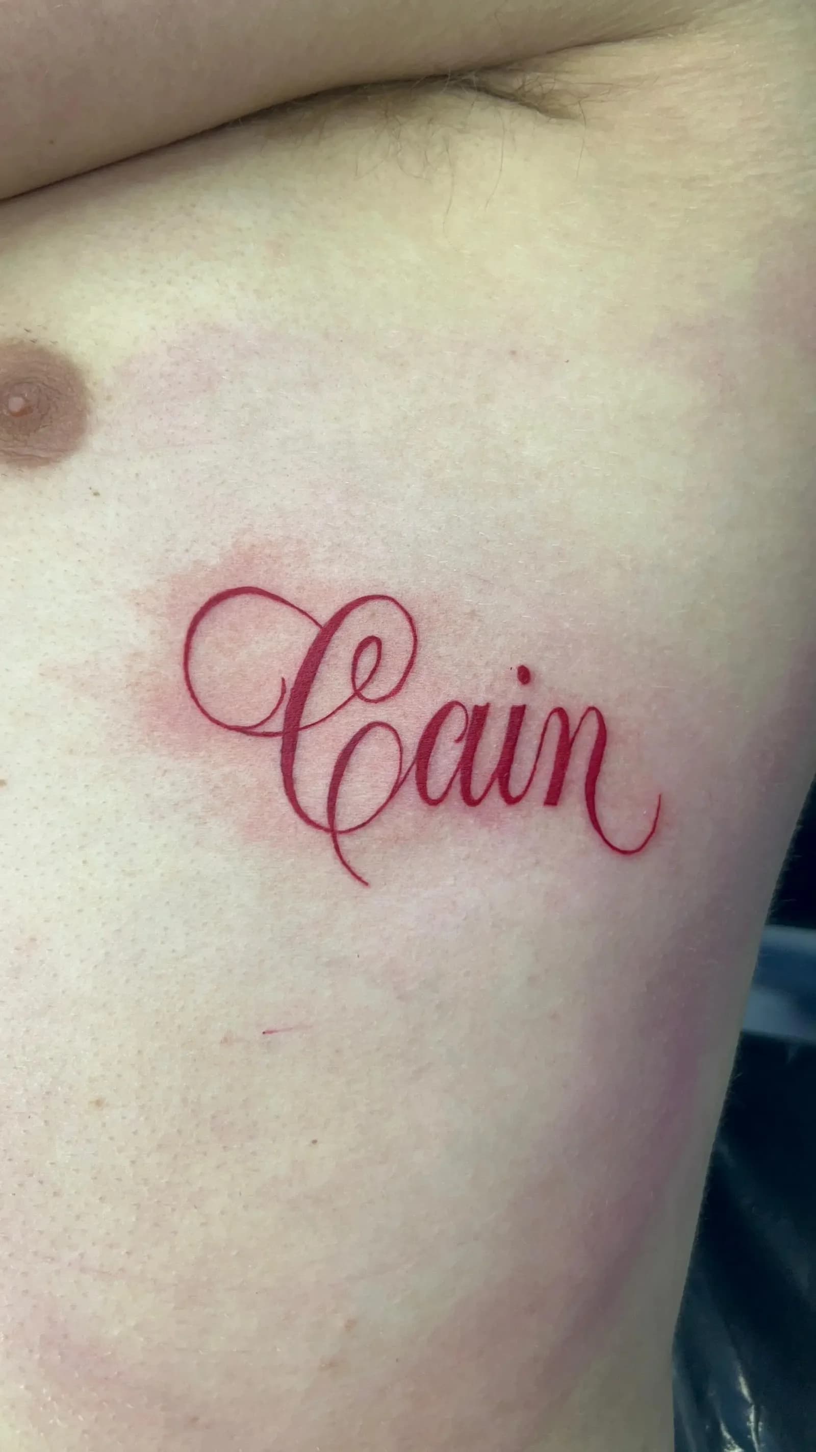 Red-ink "Cain" script tattoo on the rib cage by Gynmo at Shadow Studio Charlotte